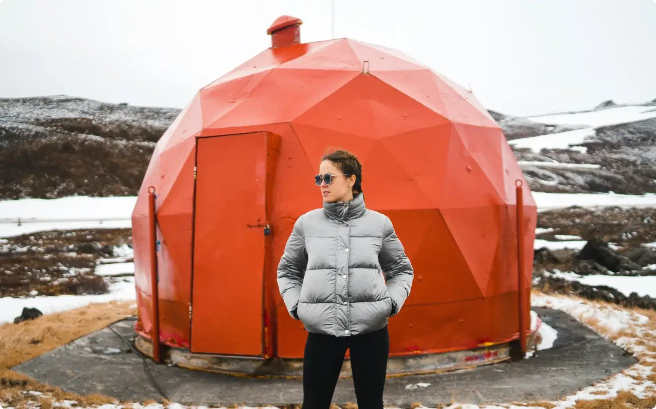 Personne en veste d'hiver se tenant devant une structure géodésique rouge unique en Islande, symbolisant l'aventure et l'opportunité de rejoindre le programme d'affiliation de Cars Iceland.