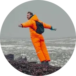 Homme en tenue d'hiver orange vif debout sur un terrain rocheux, les bras écartés.