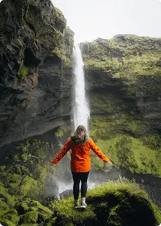 Voyageur en veste orange devant une cascade islandaise, en harmonie avec la nature et l’aventure au cœur de l’Islande.