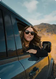 Femme penchée par la fenêtre d’une voiture avec des lunettes de soleil, profitant d’un trajet pittoresque à travers le paysage montagneux d’Islande.
