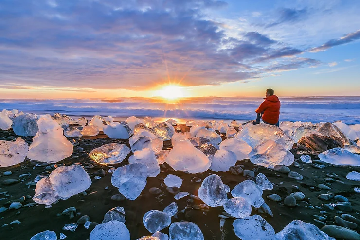 Coucher de soleil sur la plage de diamants en Islande, avec des blocs de glace scintillants et une personne en veste rouge face à l'océan.