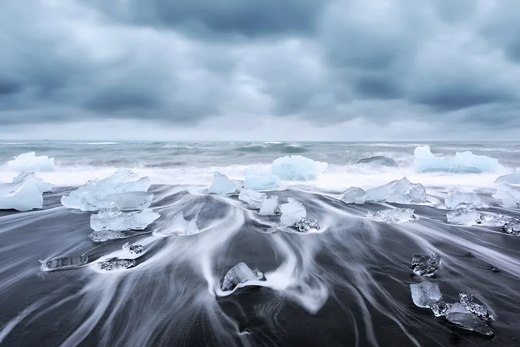 Blocs de glace sur la plage noire islandaise, avec des vagues tourbillonnantes sous un ciel nuageux dramatique.