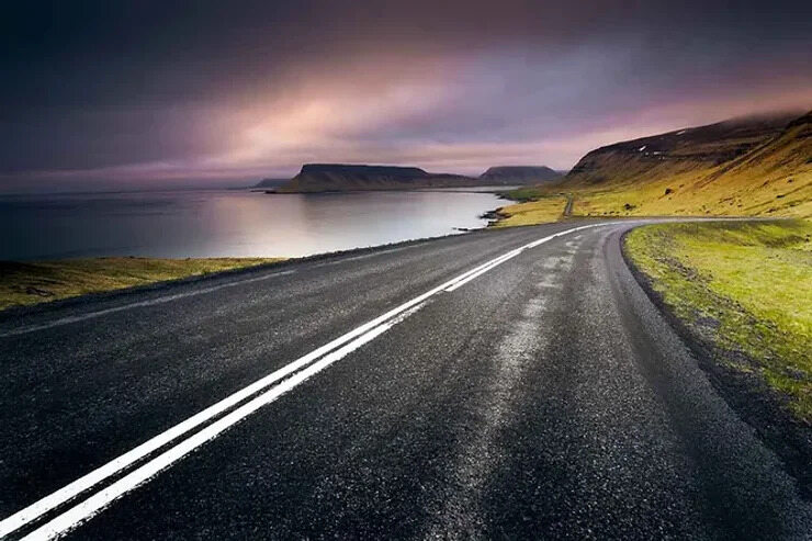 Route côtière panoramique au coucher du soleil en Islande Une autoroute islandaise côtière au crépuscule, avec un ciel dramatique aux teintes violettes et roses se reflétant sur des eaux calmes.