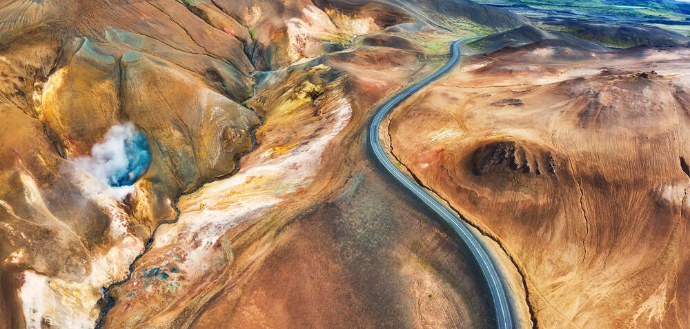 Une voiture roulant sur une route panoramique en Islande au coucher du soleil, entourée de champs verts et de montagnes au loin.
