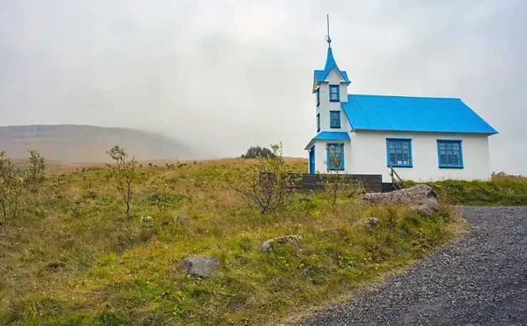 Petite église blanche avec un toit bleu vif, située dans une campagne islandaise brumeuse.