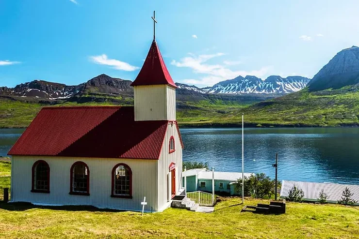 Petite église blanche avec un toit rouge surplombant un lac calme, entourée de montagnes islandaises.