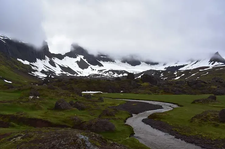 Vallée verdoyante avec un ruisseau sinueux entouré de montagnes enneigées et de nuages bas.