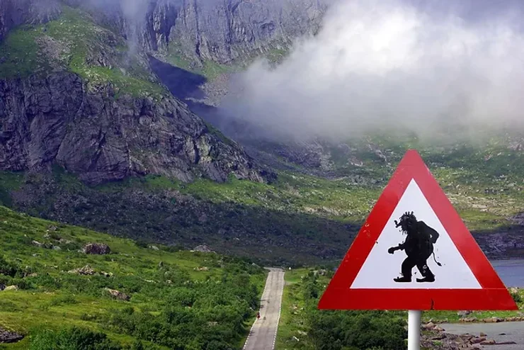 Panneau de signalisation humoristique représentant un troll marchant, situé au bord d'une route islandaise avec des montagnes en arrière-plan.