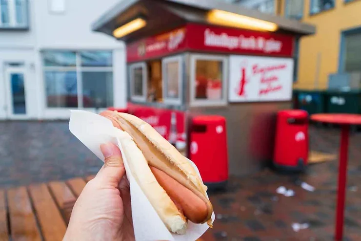 Un hot dog islandais classique avec moutarde et relish sur un petit pain, présenté devant un kiosque emblématique de Reykjavik.