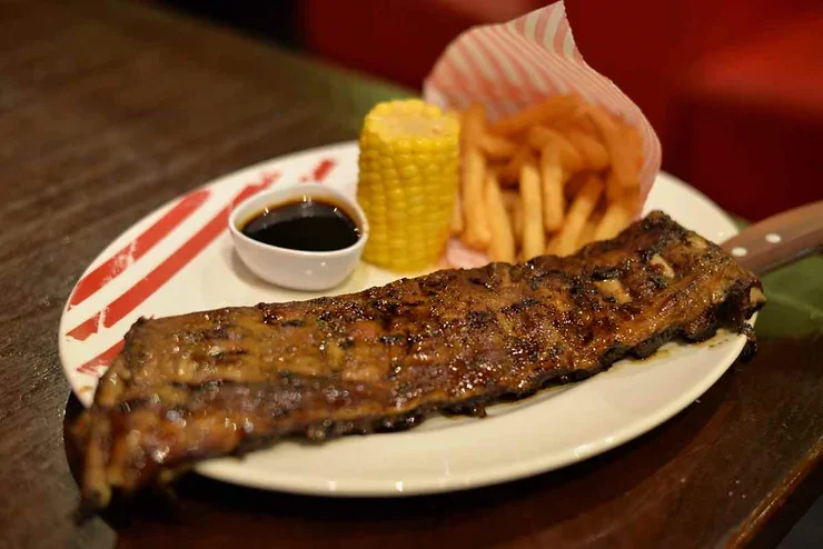 Côtes de porc grillées avec une sauce barbecue, accompagnées de frites et de maïs, servies dans un restaurant chaleureux.