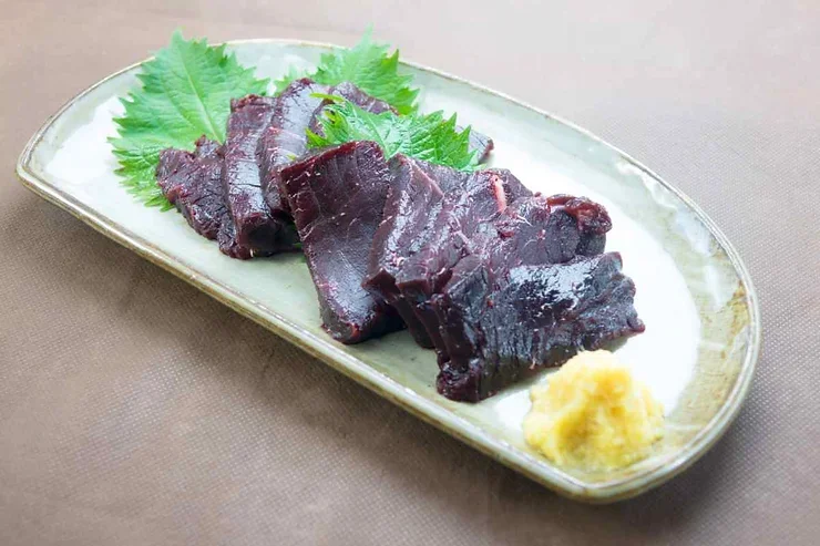 Viande de baleine Tranches de viande de baleine islandaise disposées sur un plat élégant, accompagnées de feuilles vertes fraîches et d'une petite portion de condiment jaune pour une touche de saveur traditionnelle.