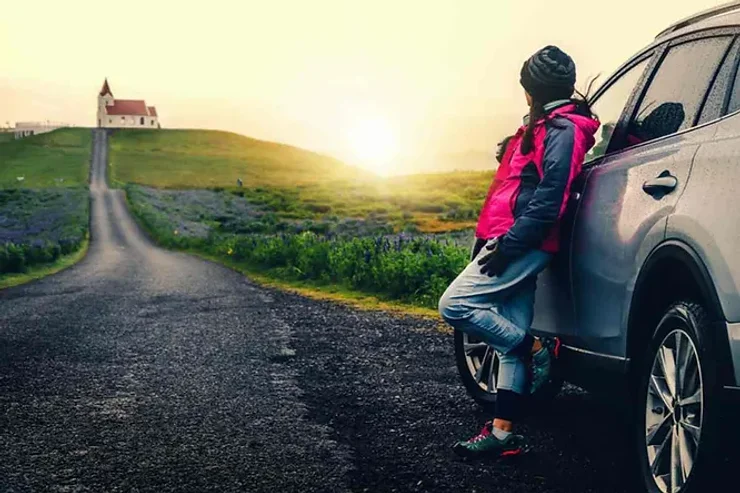 Une femme appuyée contre une voiture, regardant un coucher de soleil éclatant, avec une petite église perchée sur une colline au loin. La route qui mène à l'église est bordée de prairies verdoyantes.