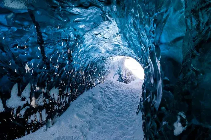 L'intérieur d'une caverne glacée avec des parois d'un bleu éclatant, sculptées par la nature, et un sol enneigé qui mène vers une ouverture lumineuse.