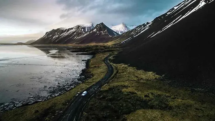 Une voiture blanche sur une route sinueuse bordée de montagnes imposantes et d'un lac calme, capturant la beauté sauvage de l'Islande.