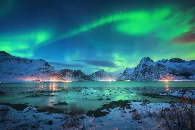 Un ciel illuminé d'aurores boréales vertes et bleues, surplombant un lac gelé entouré de montagnes enneigées. Les lumières scintillantes des villages lointains ajoutent une touche chaleureuse.