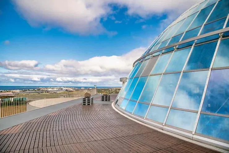 Un dôme architectural moderne en verre sous un ciel bleu parsemé de nuages blancs. La terrasse en bois qui l'entoure offre une vue panoramique sur un paysage islandais.