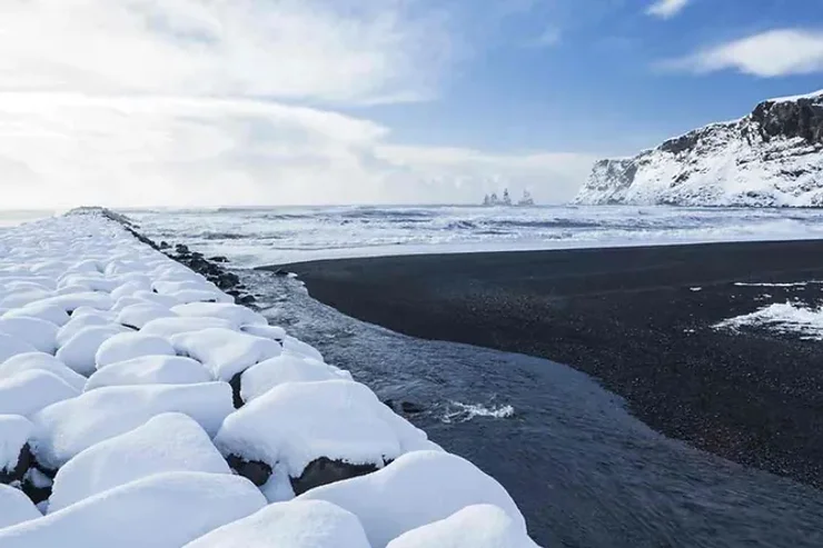 Une plage islandaise unique avec du sable noir contrastant avec la neige blanche, bordée par un océan agité et des formations rocheuses au loin.