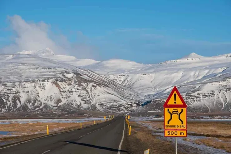 Une route islandaise bordée de montagnes enneigées, avec un panneau de signalisation jaune indiquant une 