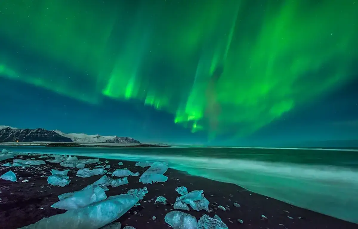 Une plage de sable noir avec des morceaux de glace scintillante sous des aurores boréales vertes et vibrantes.