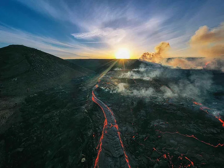 Un paysage volcanique islandais au coucher du soleil, avec une coulée de lave rougeoyante et des fumerolles qui montent dans le ciel clair.