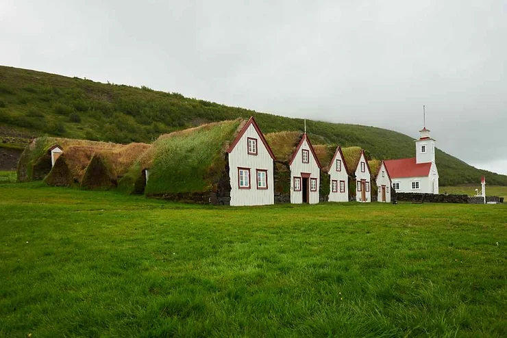 Un ensemble de maisons traditionnelles islandaises aux toits recouverts de gazon vert, entourées de collines verdoyantes et d'une petite église blanche avec un clocher rouge.