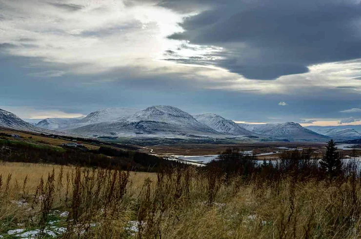 Un paysage époustouflant de montagnes enneigées sous un ciel nuageux, avec une vaste plaine et une rivière en contrebas.