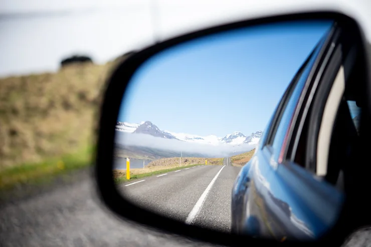 Un rétroviseur de voiture reflète une route droite menant à des montagnes enneigées sous un ciel dégagé.
