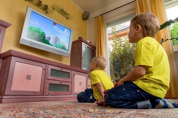 Deux jeunes garçons en t-shirts jaunes regardant attentivement un dessin animé à la télévision dans un salon chaleureux avec des rideaux jaunes et un mobilier en bois sculpté.