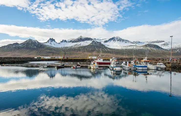 Une vue magnifique d'un petit port en Islande, avec des bateaux de pêche colorés reflétés dans une eau calme, entouré de montagnes enneigées.