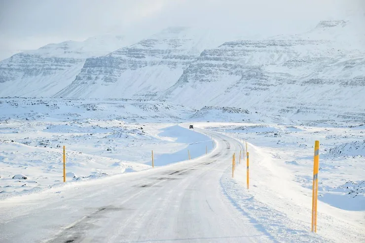 Une route isolée serpentant à travers un paysage enneigé spectaculaire en Islande, bordée de piquets jaunes pour guider les conducteurs.