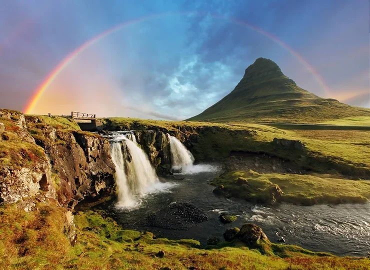 Une image captivante du mont Kirkjufell, accompagné de sa cascade voisine et d'un arc-en-ciel vibrant qui illumine le ciel.