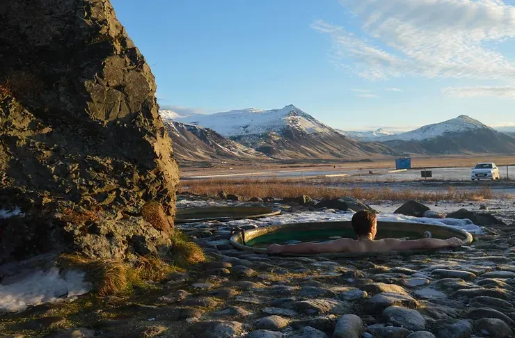 Une source chaude naturelle entourée de montagnes enneigées, où une personne se relaxe en profitant du paysage hivernal islandais.