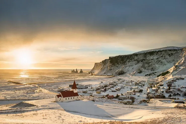 L'église emblématique de Vik se dressant fièrement surplombant un village enneigé, avec un coucher de soleil éclatant en arrière-plan.
