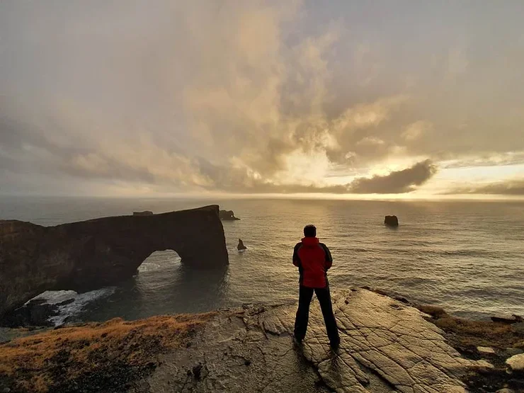 Une silhouette d'une personne contemplant l'arche rocheuse majestueuse de Dyrhólaey, baignée par la lumière douce du coucher de soleil.