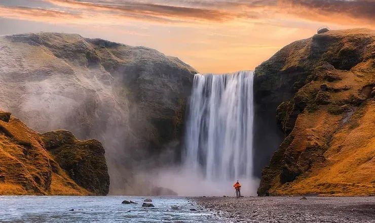 La cascade de Skógafoss illuminée par une lumière dorée, entourée d'une végétation luxuriante et de collines imposantes.