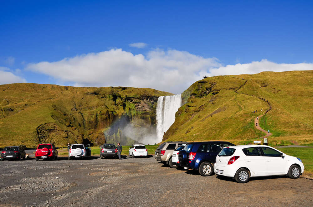 Un parking avec plusieurs voitures devant la majestueuse cascade de Skógafoss, entourée de collines verdoyantes et d’un ciel bleu avec quelques nuages légers.
