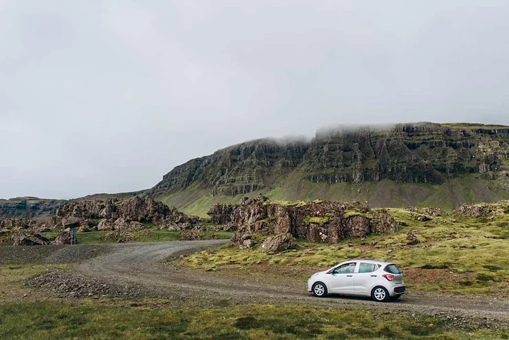 Une petite voiture blanche sur une route de gravier dans un paysage islandais. Des collines rocheuses et une montagne brumeuse se trouvent à l'arrière-plan.