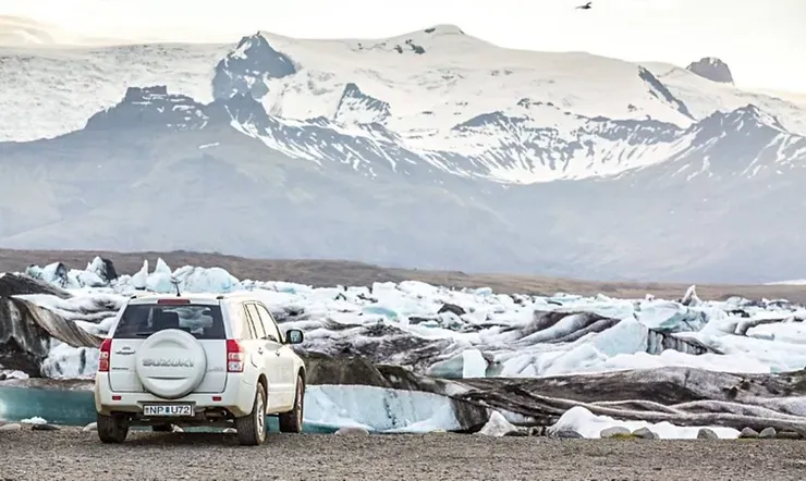 Un SUV blanc garé devant un paysage de glacier en Islande. Les montagnes enneigées se reflètent dans les eaux glacées.