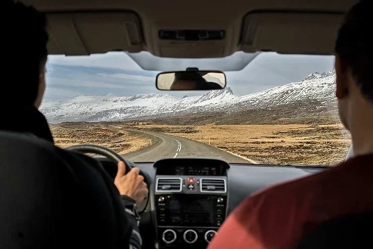 Vue depuis l'intérieur d'une voiture avec deux personnes à bord, conduisant sur une route sinueuse entourée de plaines et de montagnes enneigées. Le ciel est clair.
