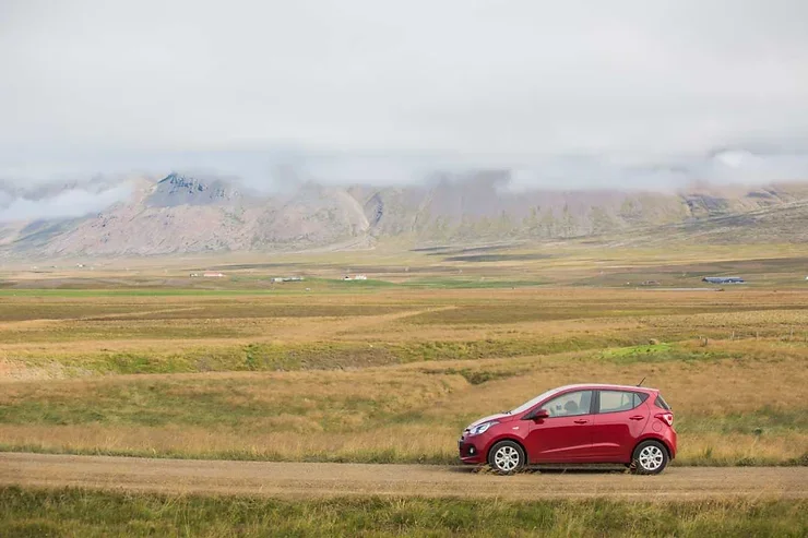 Une petite voiture rouge sur une route de gravier, entourée de vastes prairies et de montagnes brumeuses à l'arrière-plan.