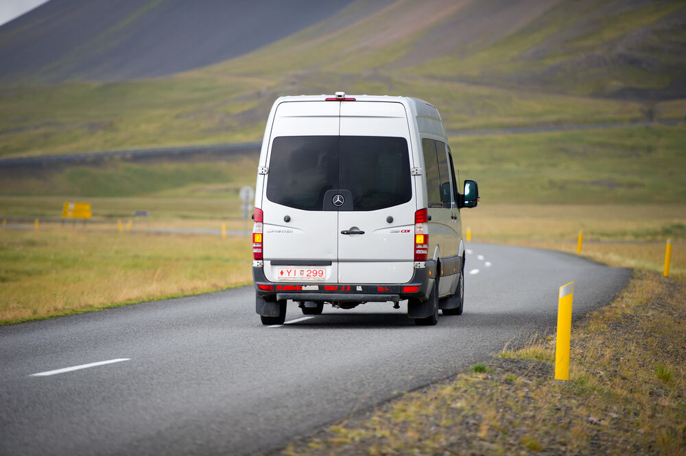 Une camionnette blanche roulant sur une route asphaltée dans une vallée islandaise, entourée de collines vertes sous un ciel couvert.