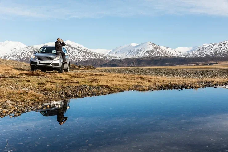Un SUV gris argenté garé près d'un lac paisible, avec des montagnes enneigées en arrière-plan. Une personne prend des photos depuis le côté de la voiture.