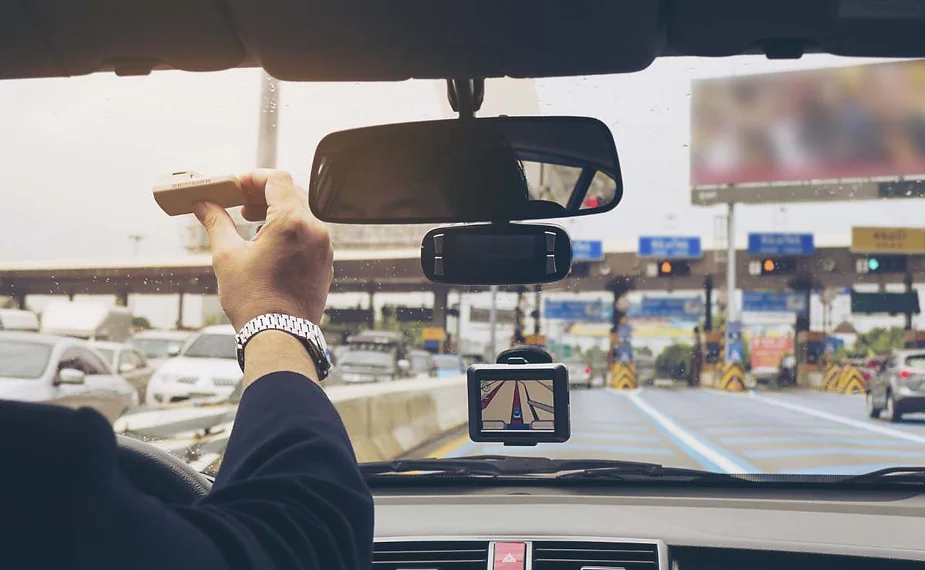 Un conducteur à l’intérieur d’une voiture tend la main vers le pare-brise avec un badge de péage, prêt à entrer dans une voie de péage ; un GPS affiche l’itinéraire sur le tableau de bord et d’autres véhicules sont visibles devant.