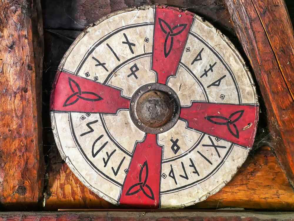 Un grand bouclier circulaire en bois dont les parties sont peintes en rouge et en blanc, décoré de runes et de symboles nordiques. Au centre se trouve un boîtier métallique rond.