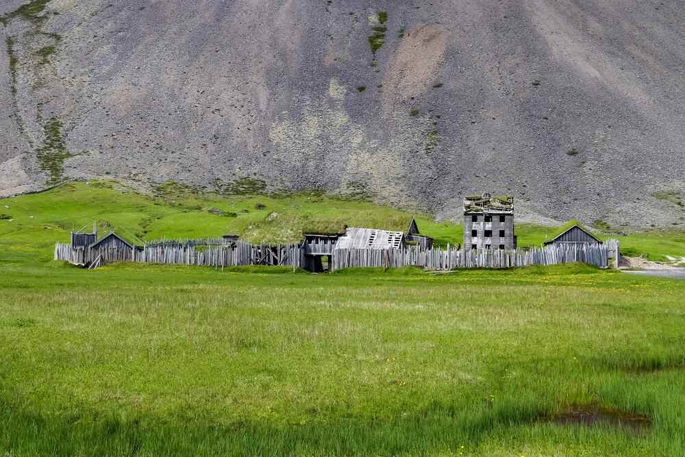 Reproduction d'un ancien village viking avec palissades et tourelles en bois, situ&eacute; dans une plaine verdoyante au pied d'une montagne grise et escarp&eacute;e.