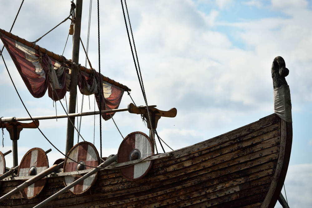 D&eacute;tail d'un bateau viking avec des boucliers en bois color&eacute;s le long des flancs et des voiles pli&eacute;es, pris avec un ciel nuageux en arri&egrave;re-plan.