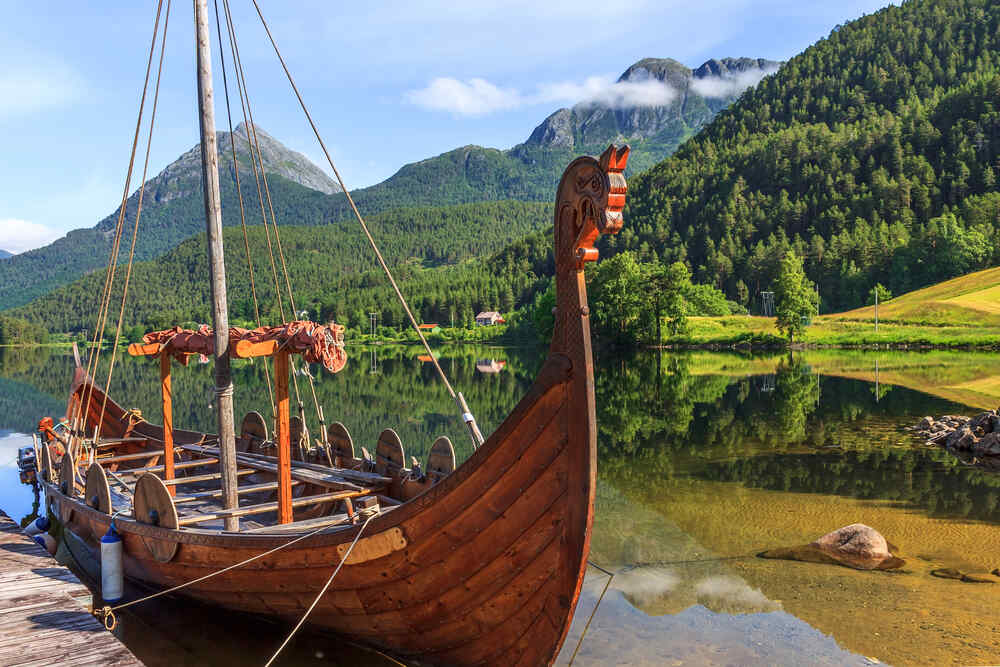 Un bateau viking en bois avec une t&ecirc;te de dragon sculpt&eacute;e dans la proue, amarr&eacute; sur un lac tranquille entour&eacute; de montagnes verdoyantes et d'un ciel clair.
