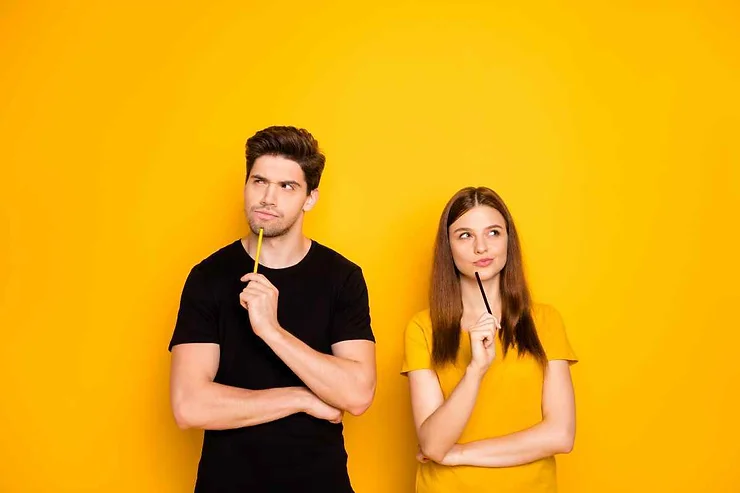Deux jeunes, un homme en noir et une femme en jaune, réfléchissent en tenant un crayon devant un fond jaune lumineux.