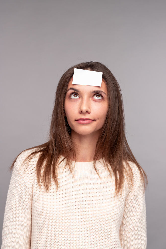 Une jeune fille vêtue d'un pull crème avec un morceau de papier blanc collé sur son front regarde vers le haut avec un air pensif, participant probablement à un jeu de devinettes.