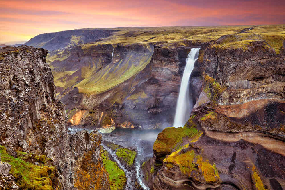 Vue panoramique de la cascade Haifoss sous un ciel aux teintes orangées, avec ses chutes impressionnantes et ses rochers moussus.
